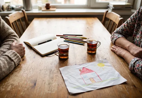 Een keukentafel met twee glazen thee, een opengeslagen schrift, kleurpotloden en een kindertekening; twee volwassenen in gesprek